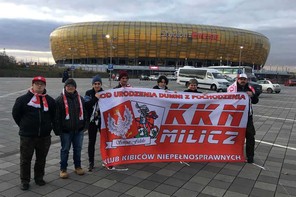 Zdjęcie wykonane wczesnym wieczorem przed stadionem w Gdańsku. Siedmiu kibiców trzyma biało-czerwoną flagę KKN MILICZ. Za nimi widoczny stadion, który swoją kolorystyką przypomina bursztyn.