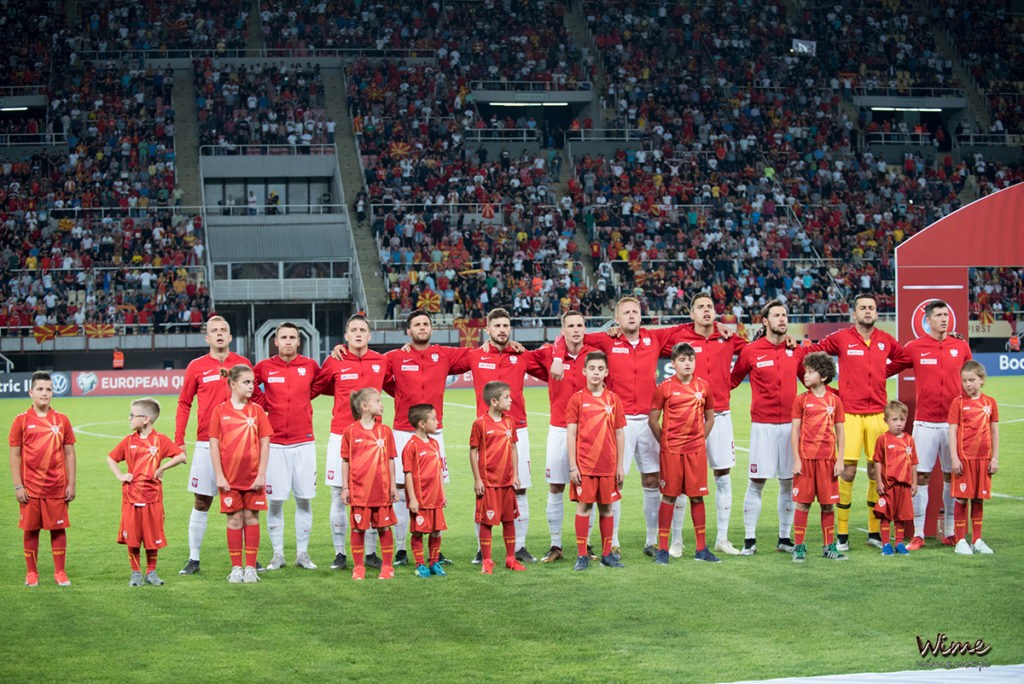 Zdjęcie kolorowe-na płycie boiska stoją w szeregu piłkarze, przed nimi stoją młodzi kibice, w oddali na sektorach stoją kibice.